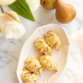 Baked cheese and prosciutto covered pears in a crescent roll on a white platter. Flowers and pears to the side.