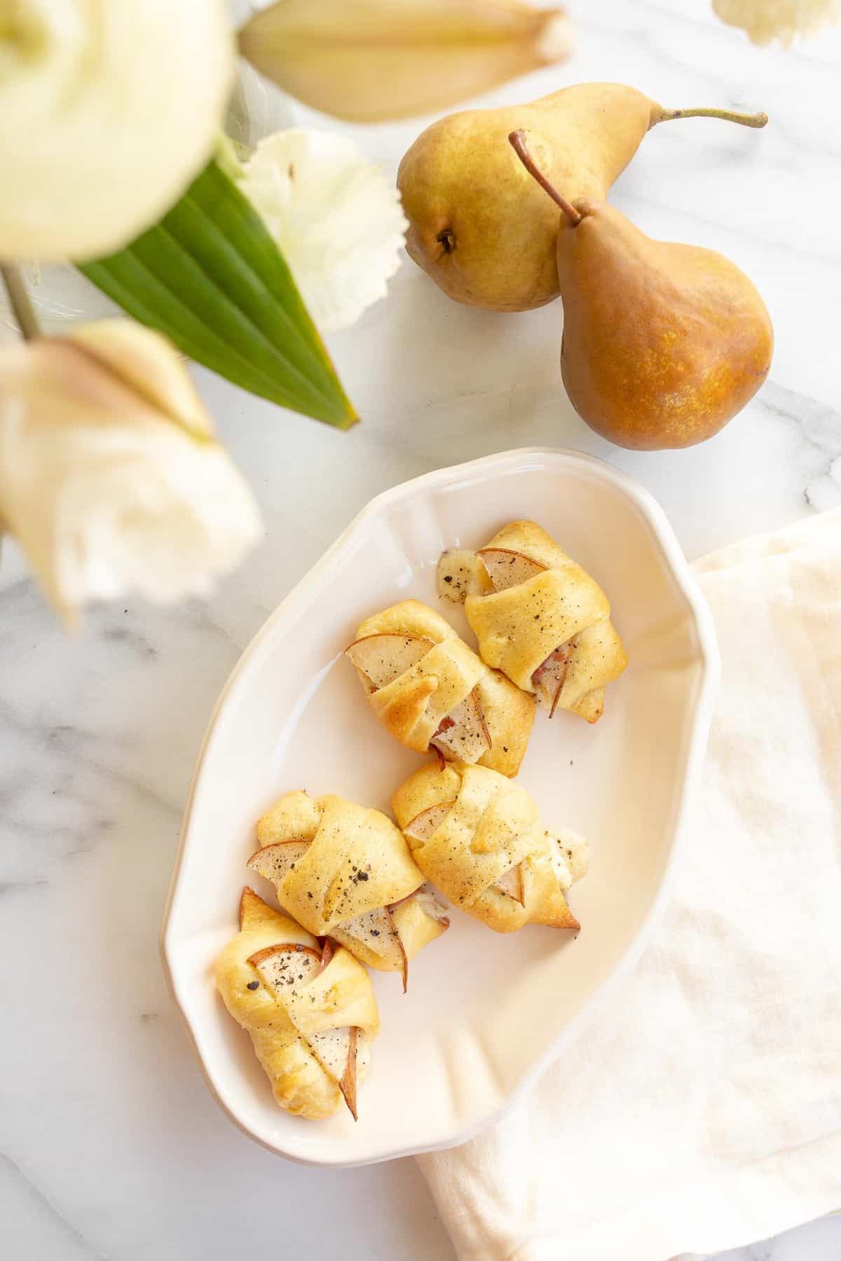 Baked cheese and prosciutto covered pears in a crescent roll on a white platter. Flowers and pears to the side. 
