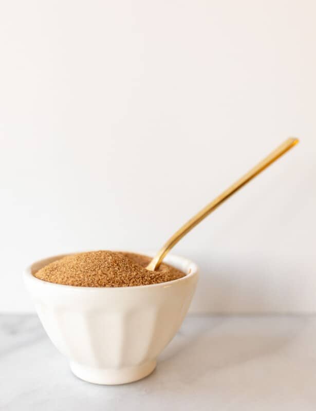 White marble surface, white ceramic bowl of cinnamon sugar recipe with gold spoon sticking up.