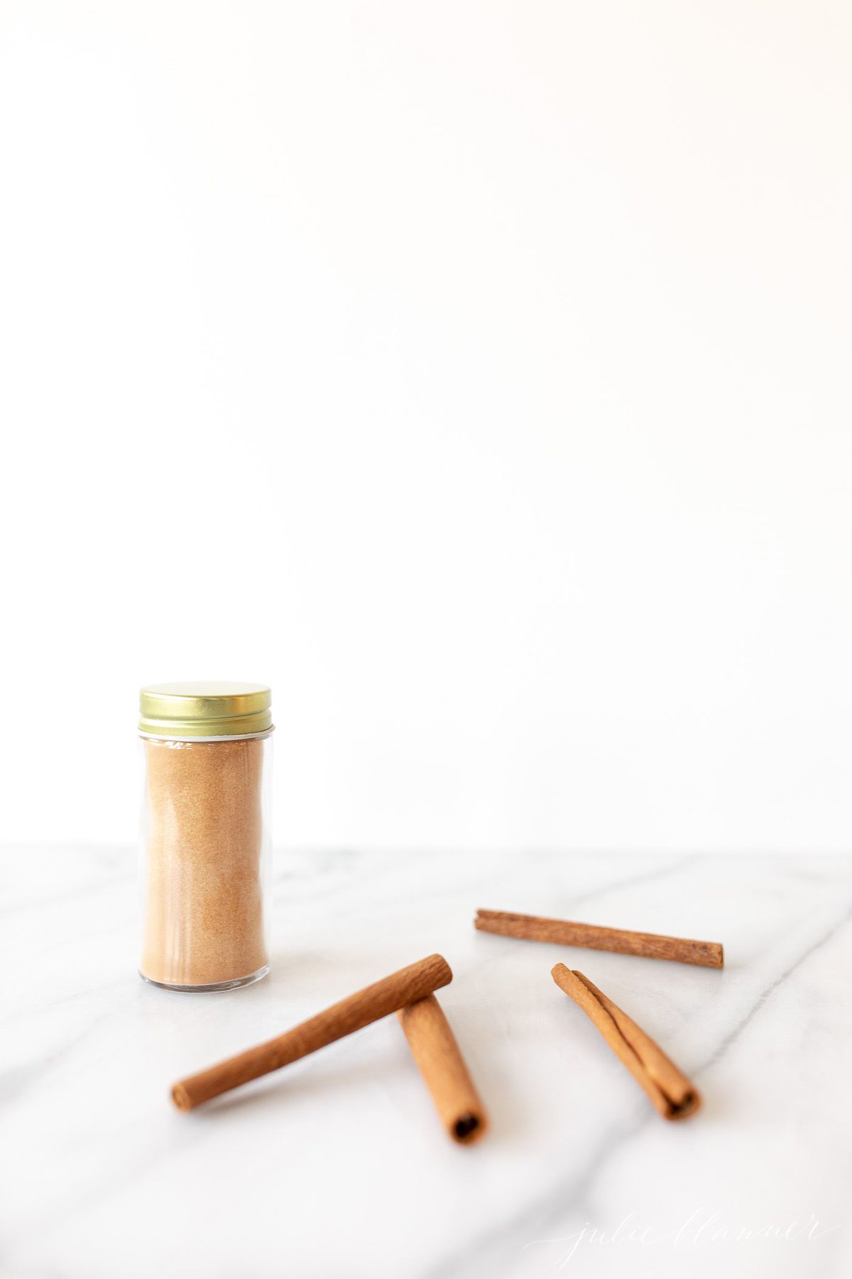 A glass jar of a cinnamon sugar recipe against a white background. Cinnamon sticks scattered around.