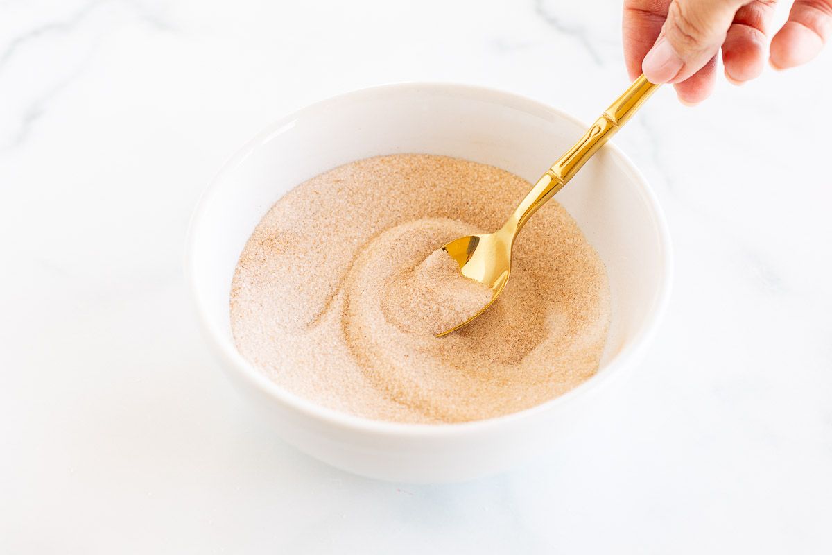 A bowl of cinnamon sugar with a hand stirring a gold spoon.