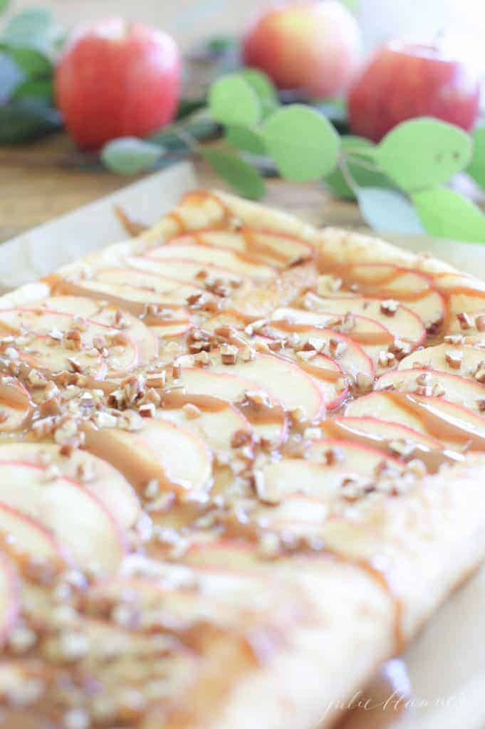 Apple tart topped with stripes of caramel and nuts, touch of greenery in background.