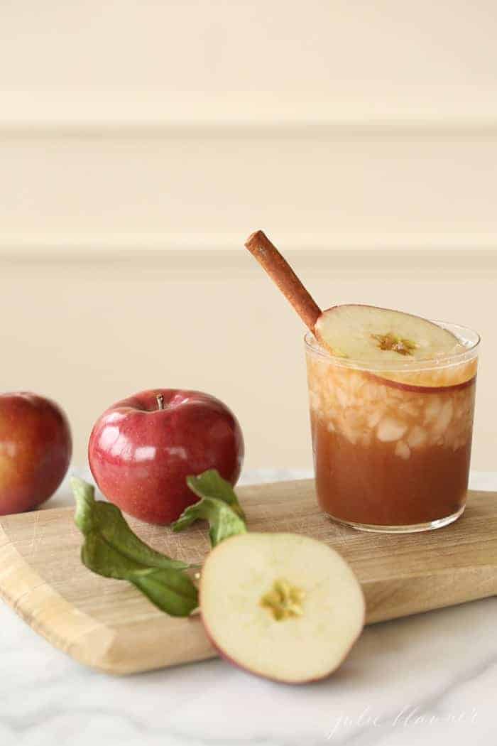 A wooden cutting board with an apple old-fashioned, garnished with a cinnamon stick. Apples are around it on the cutting board.