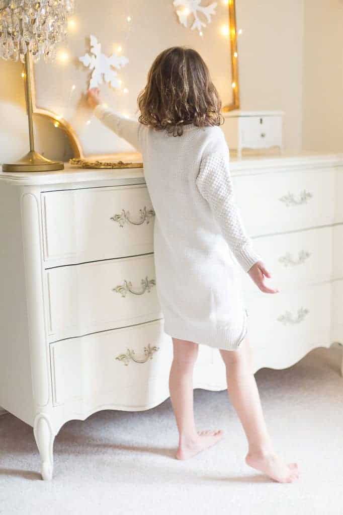 A little girl dressed in white, hanging paper snowflakes for simple Christmas decor in her white bedroom.