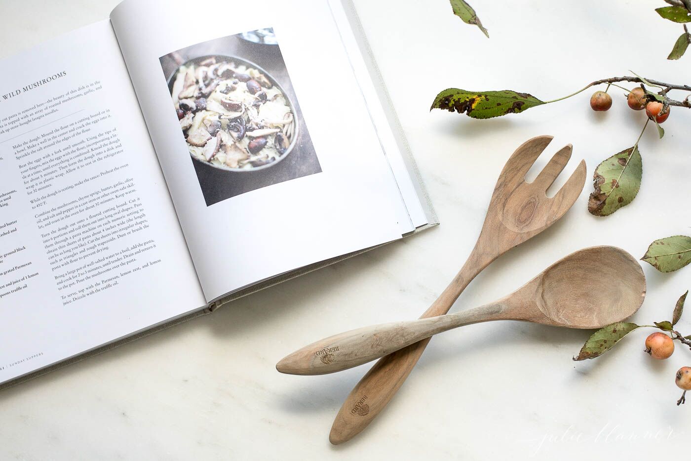 Marble surface, open cookbook, fall berries and wooden spoons to the side.