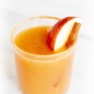 Cinnamon liquor cocktail glass on marble surface, garnished with apple slice and cinnamon sticks.