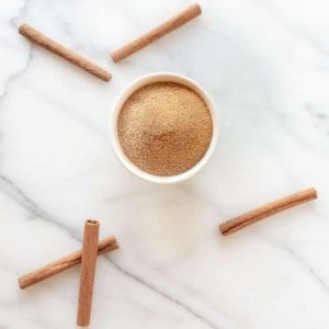 A bowl of cinnamon sugar on a marble surface, surrounded by cinnamon sticks.