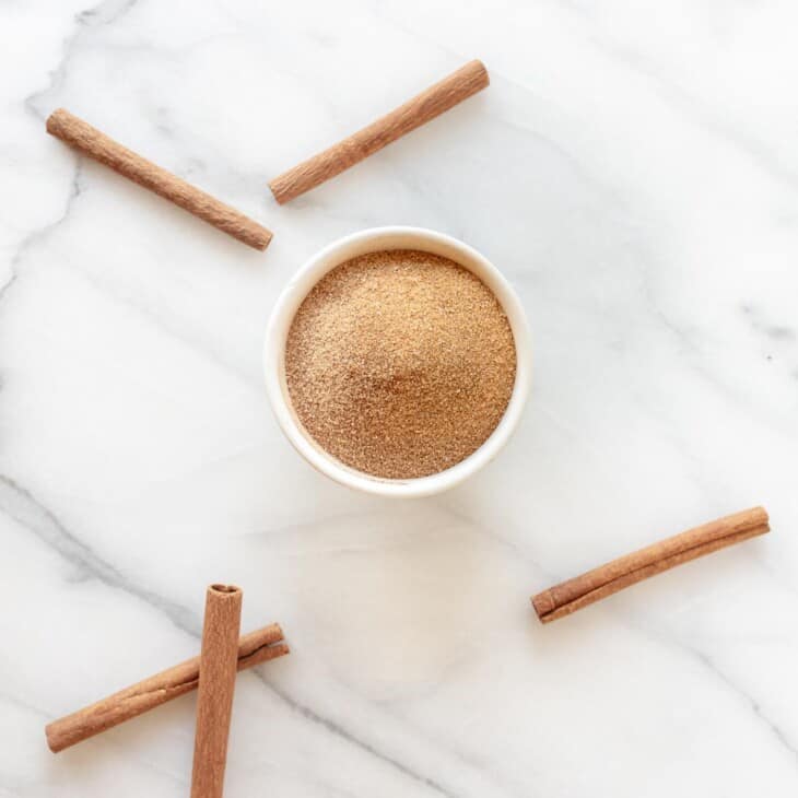 Marble surface with a bowl of the perfect cinnamon sugar ratio, cinnamon sticks strewn about.