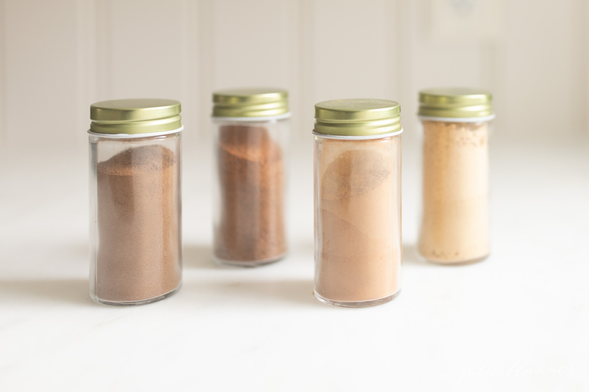 Four glass spice jars with gold lids on a white kitchen countertop.