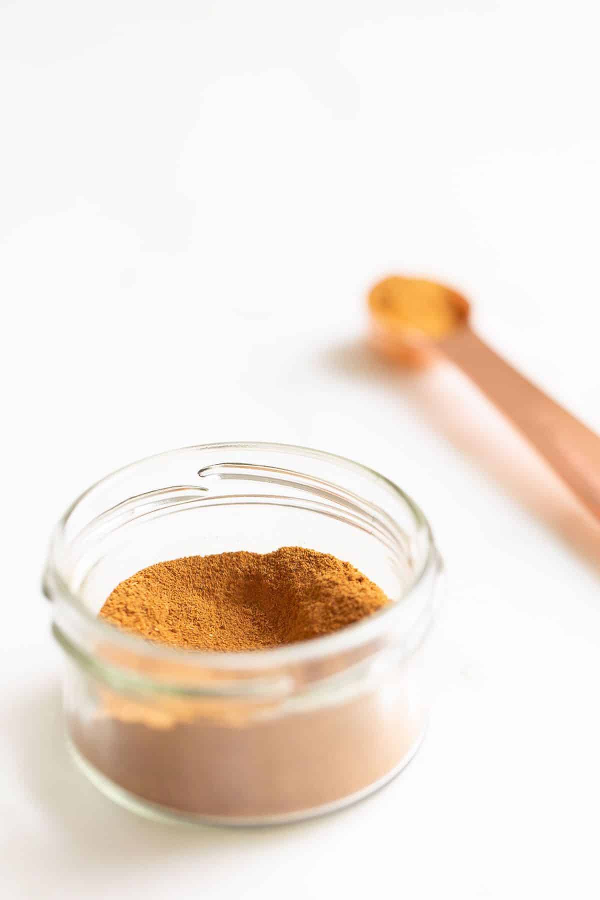 Small glass jar filled with spice mix on a white surface, teaspoon in background.