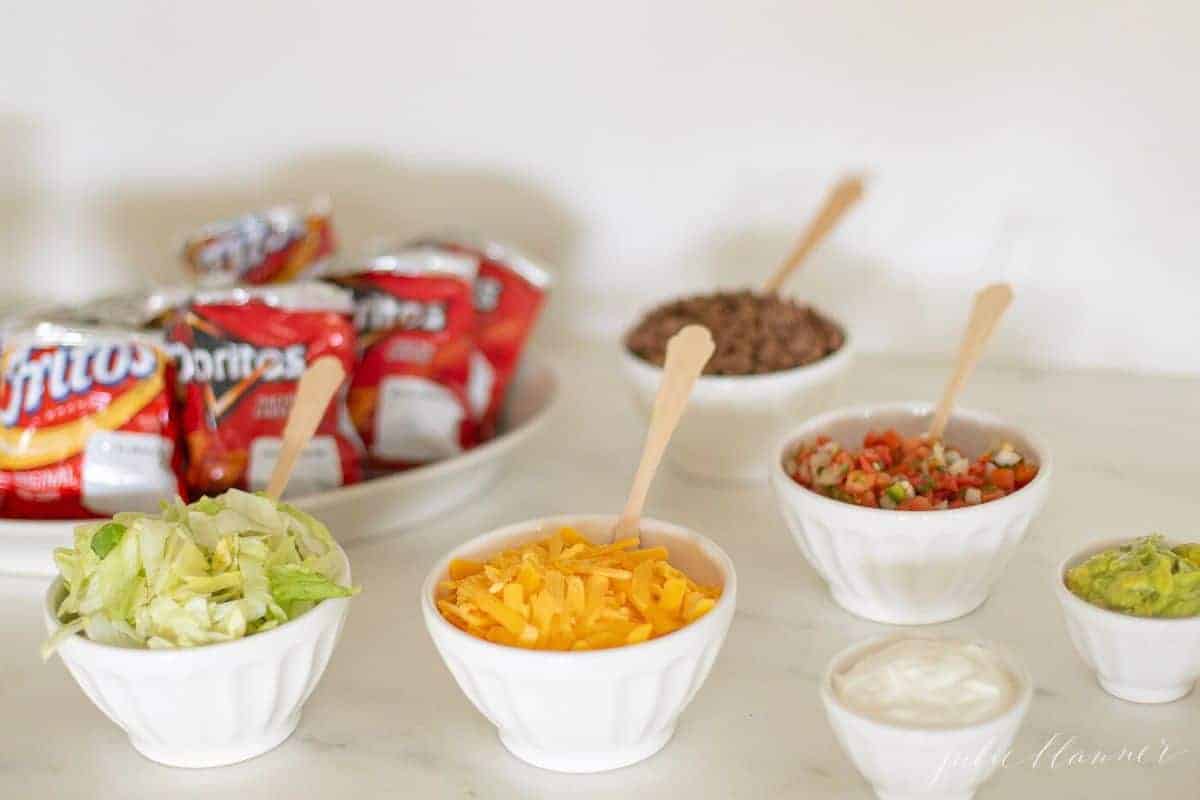 Walking taco bar display with chips and various toppings in white bowls.