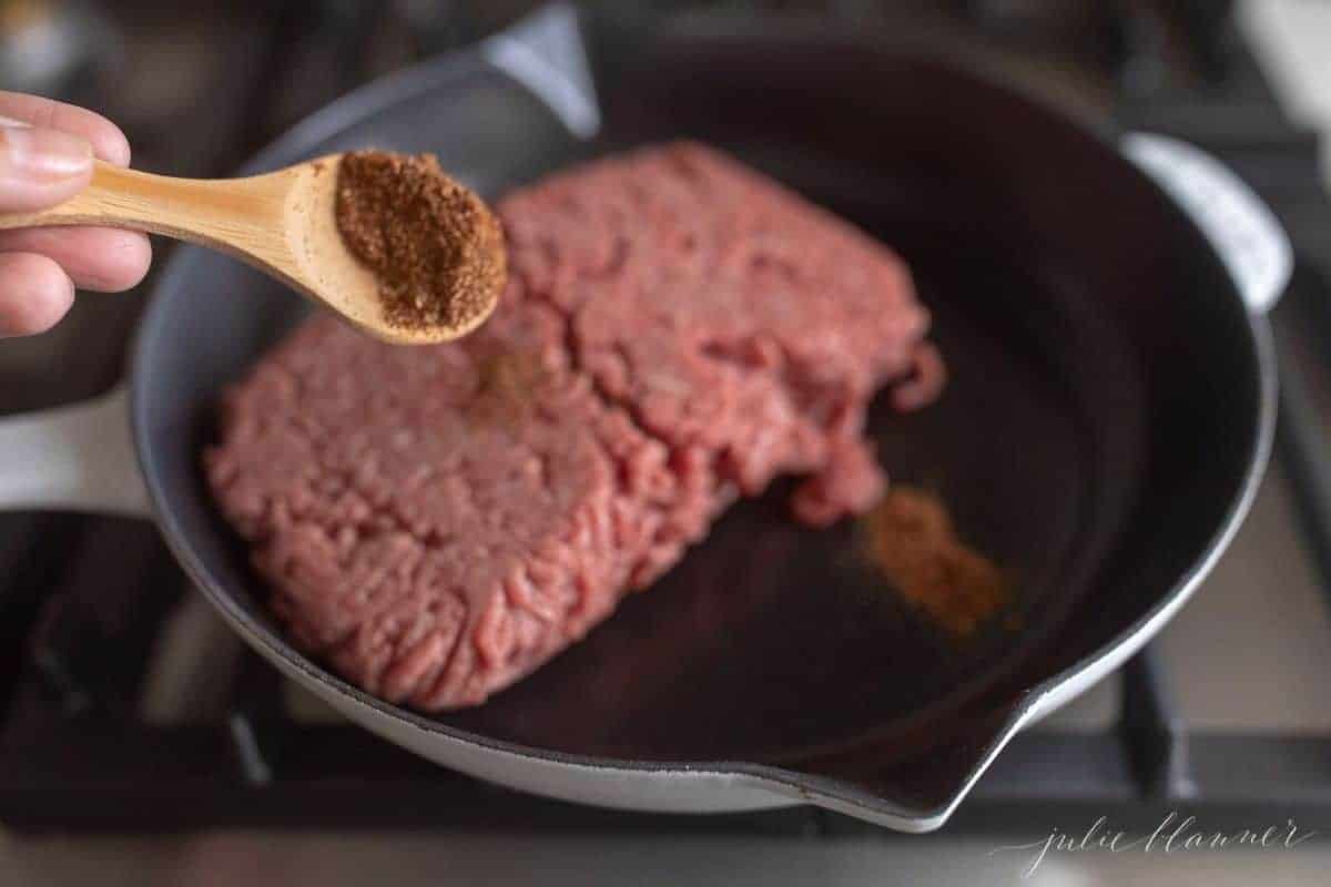 Skillet with a block of raw ground beef, wooden spoon reaching in with seasonings.