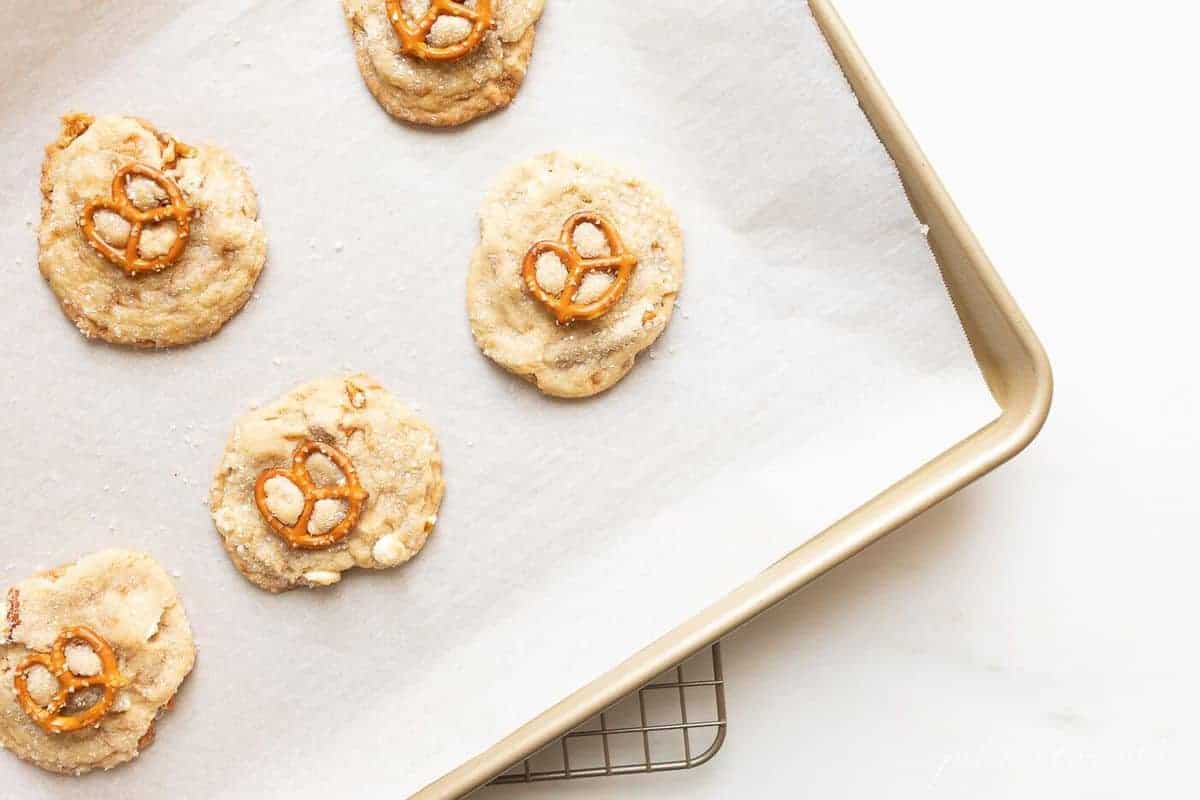 Gold baking sheet lined with parchment paper, white chocolate chip cookies topped with pretzel on top.
