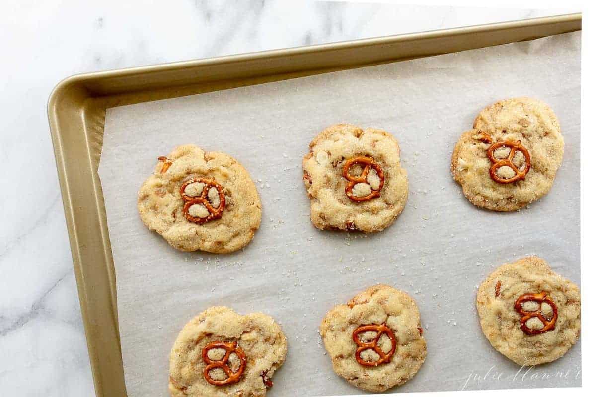 Gold baking sheet lined with parchment paper, white chocolate chip cookies topped with pretzel on top.