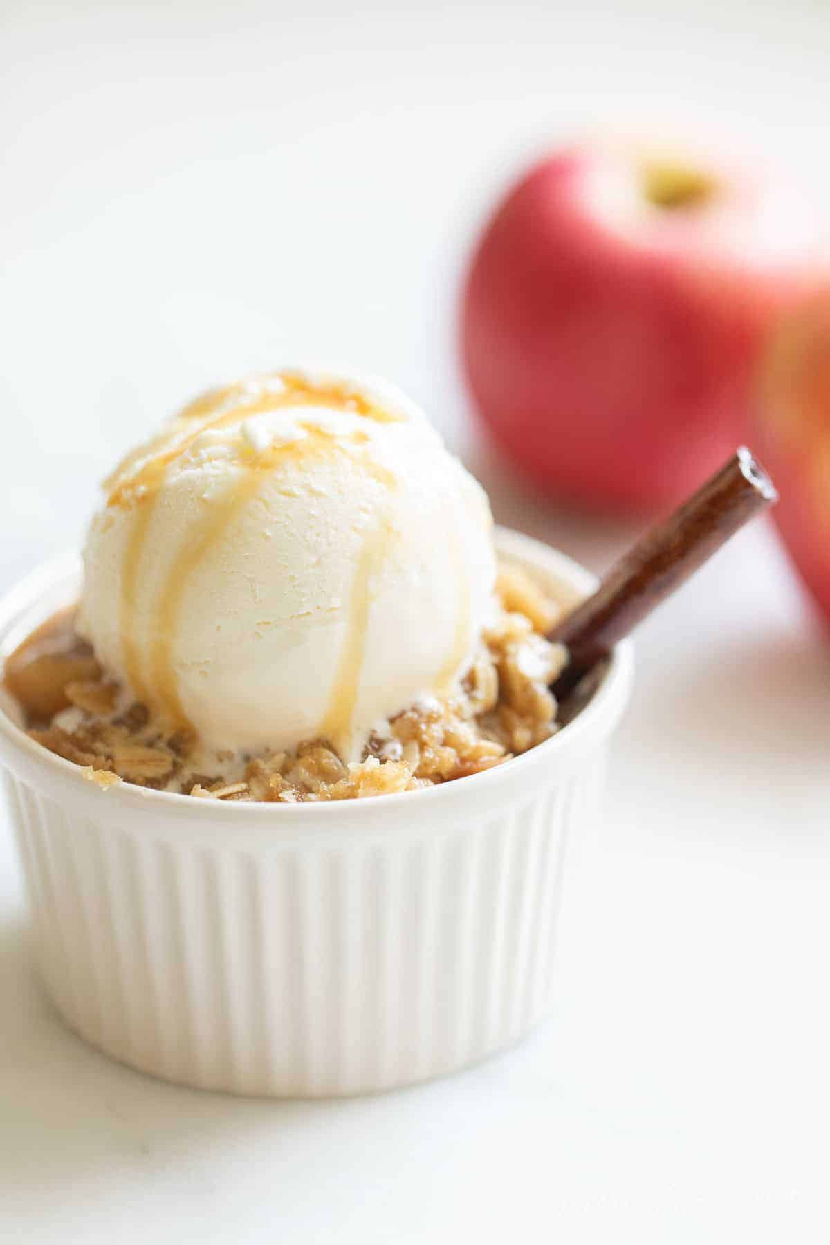 White surface with a small white ramiken of apple crisp, scoop of vanilla ice cream and caramel on top. apples in the background.
