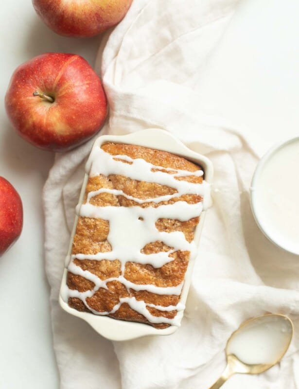 A white surface, apples and frosting to the side, with a loaf of apple bread, glaze drizzled on top.