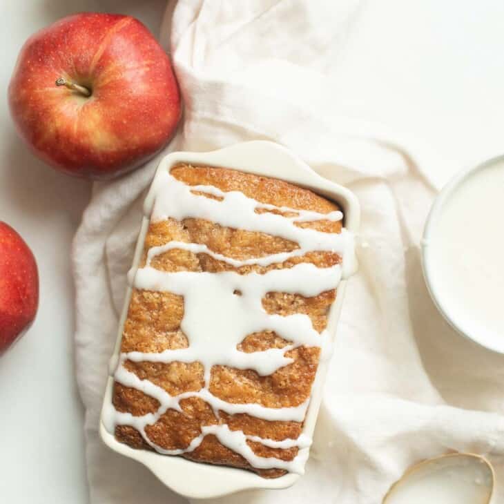 A white surface, apples and frosting to the side, with a loaf of apple bread, glaze drizzled on top.
