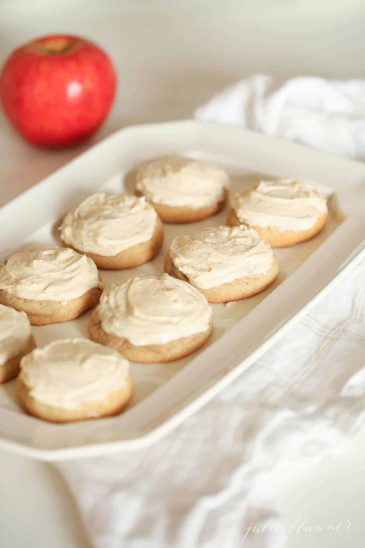 White tray on top of white linen towel, apple in background- frosted apple sugar cookies on tray.
