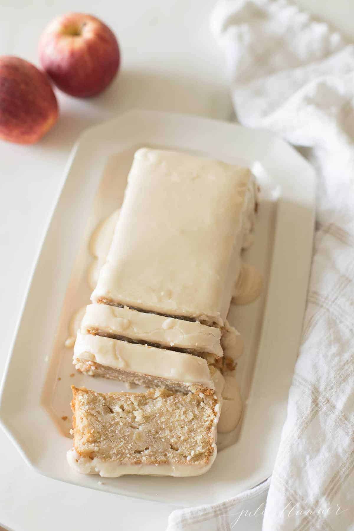 A white rectangular tray, slices of a pound cake recipe covered in a glaze, apples in background.