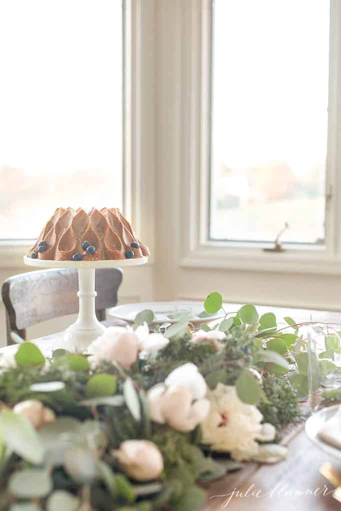A dining table with flowers, and a bundt cake on a tray to the side.