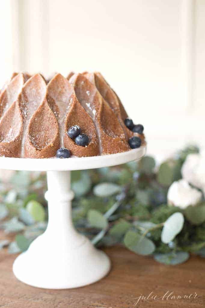 A dining table with flowers, and a bundt cake with blueberries on a tray to the side.