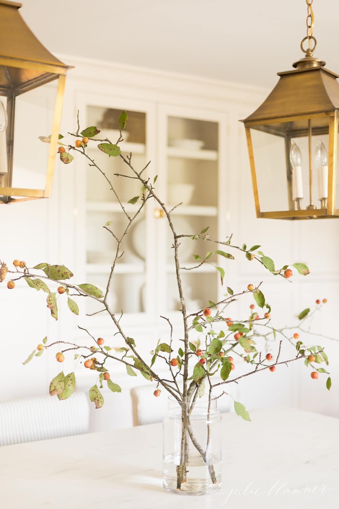 Fall branches with berries in a glass vase, in an all white kitchen, brass lanterns above.