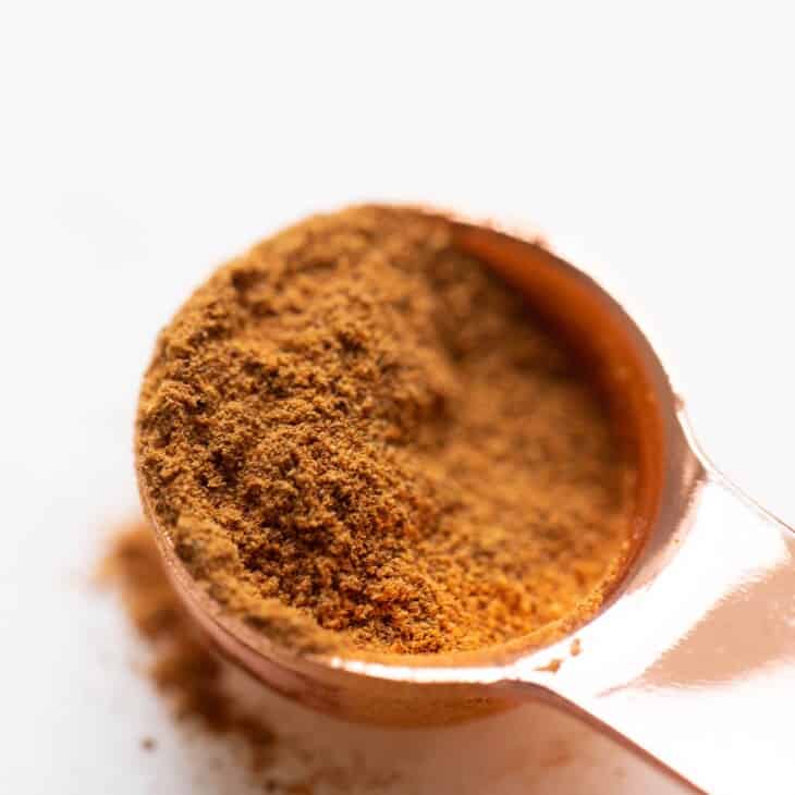 White surface, copper tablespoon filled with pumpkin pie spice mix, loose spices on surface.