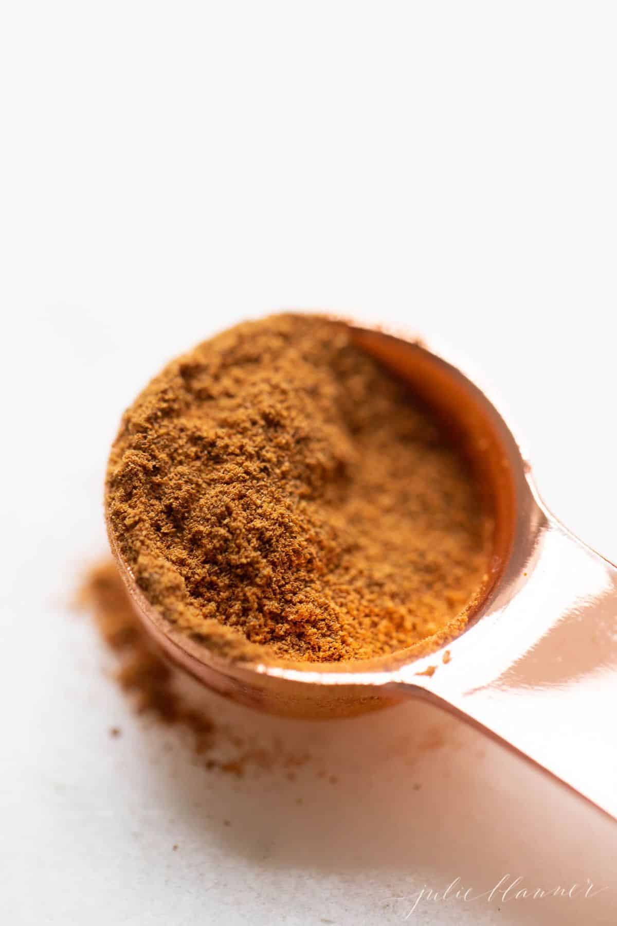 White surface, copper tablespoon filled with pumpkin pie spice mix, loose spices on surface.