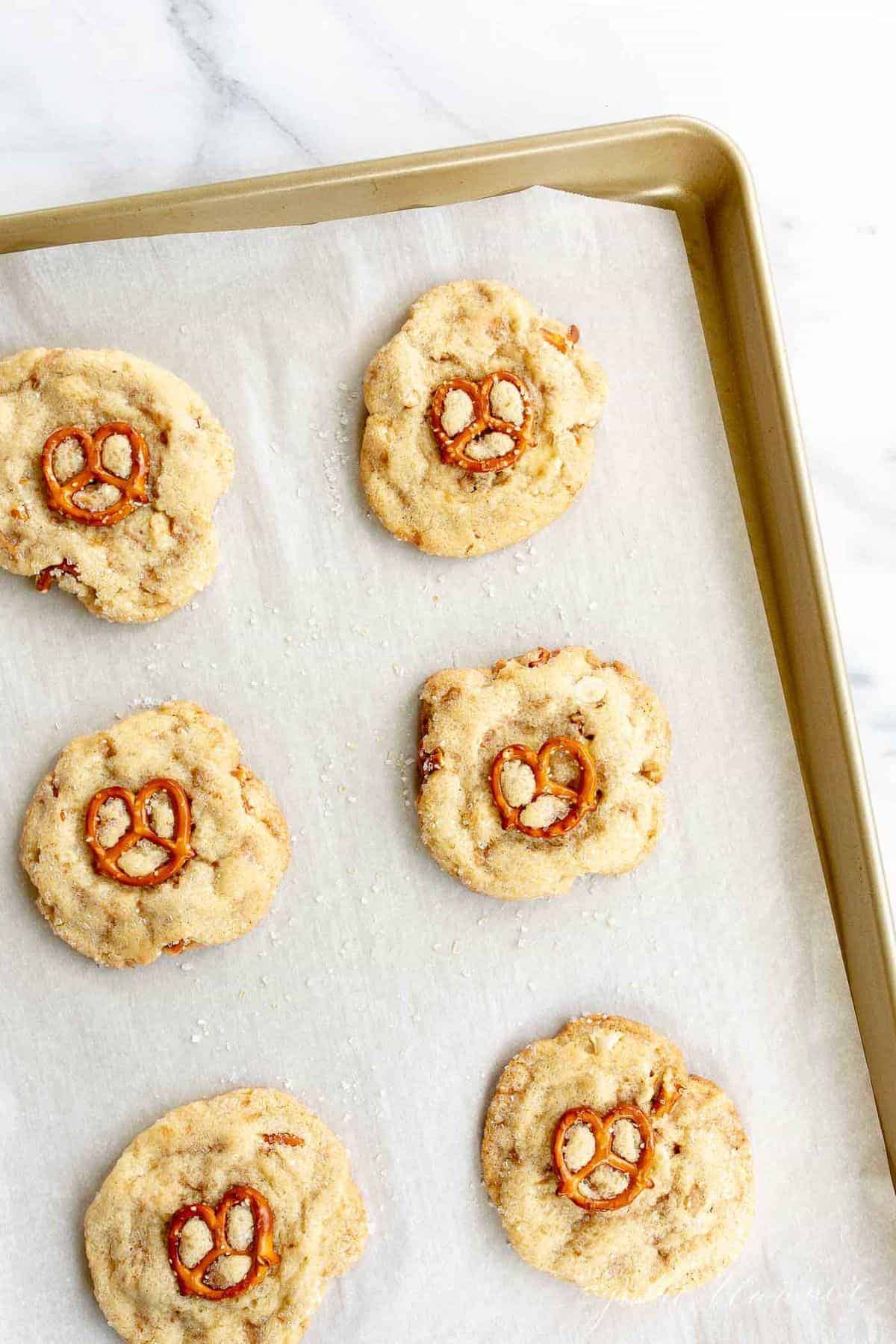 Gold baking sheet lined with parchment paper, white chocolate chip cookies topped with pretzel on top.