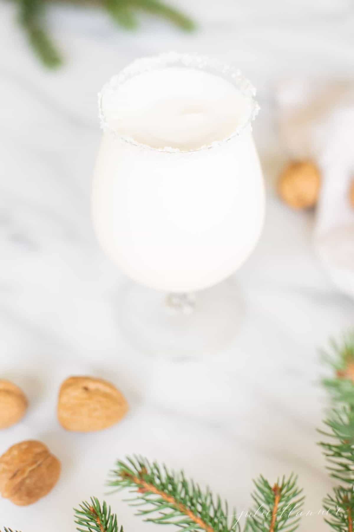 Marble surface with a clear glass filled with a snowball drink, rimmed in sugar, with chestnuts and evergreen touches around.