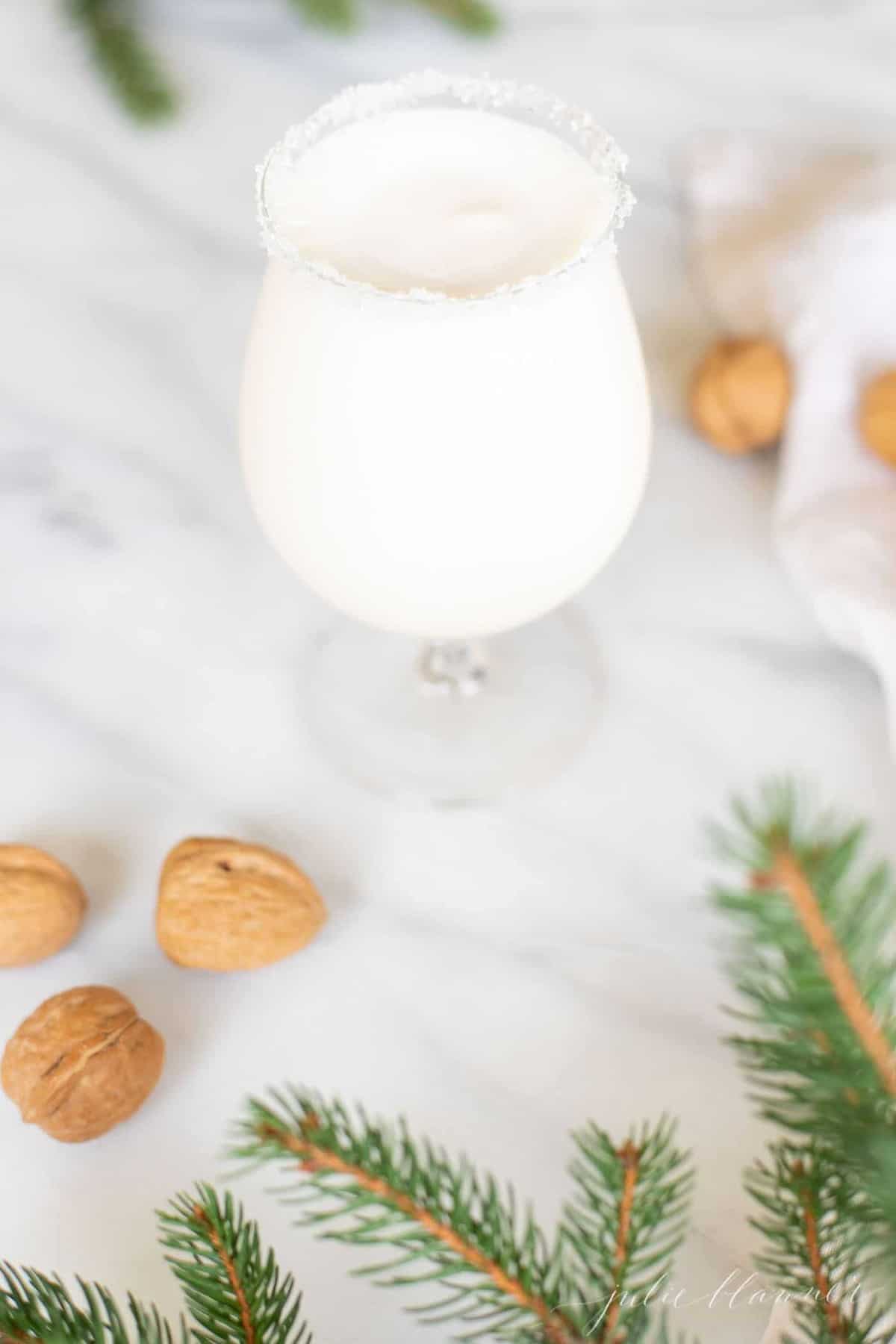 Marble surface with a clear glass filled with a snowball drink, rimmed in sugar, with chestnuts and evergreen touches around.