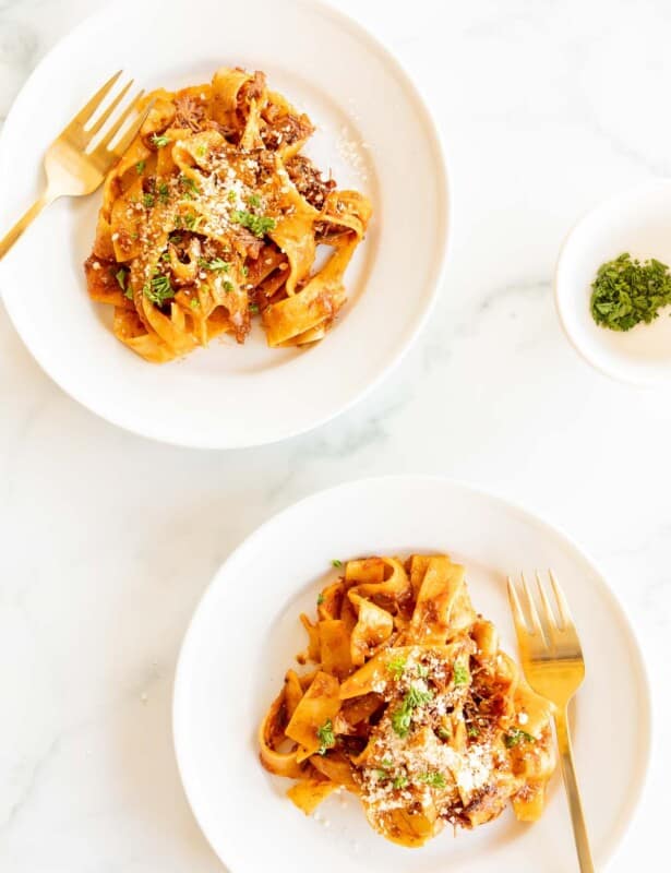 Two white plates on a marble surface, filled with a beef ragu recipe and a small bowl of parsley to the side.