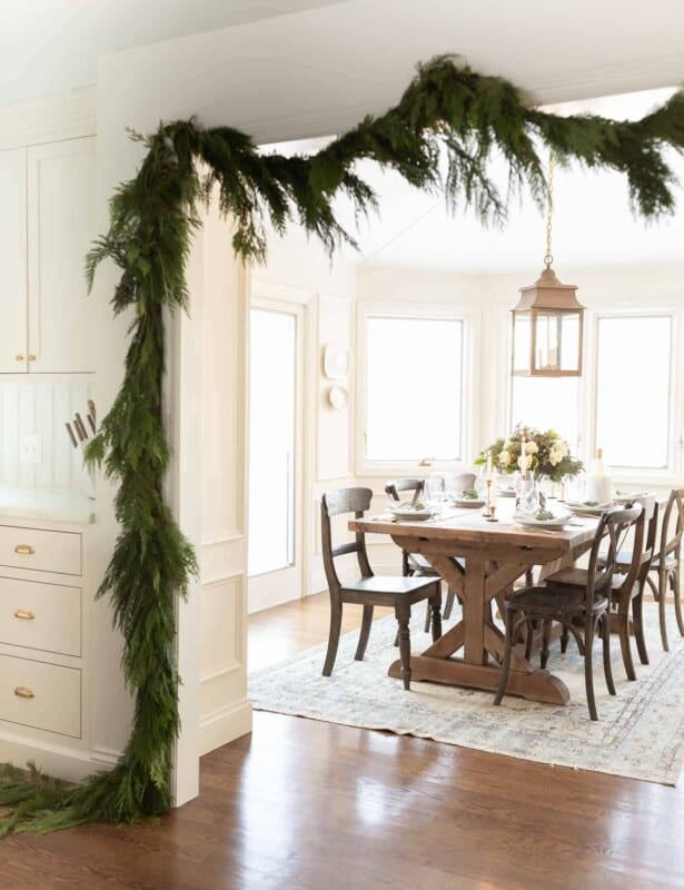 traditional kitchen draped in cedar garland peeking into breakfast nook with blue christmas flower arrangement