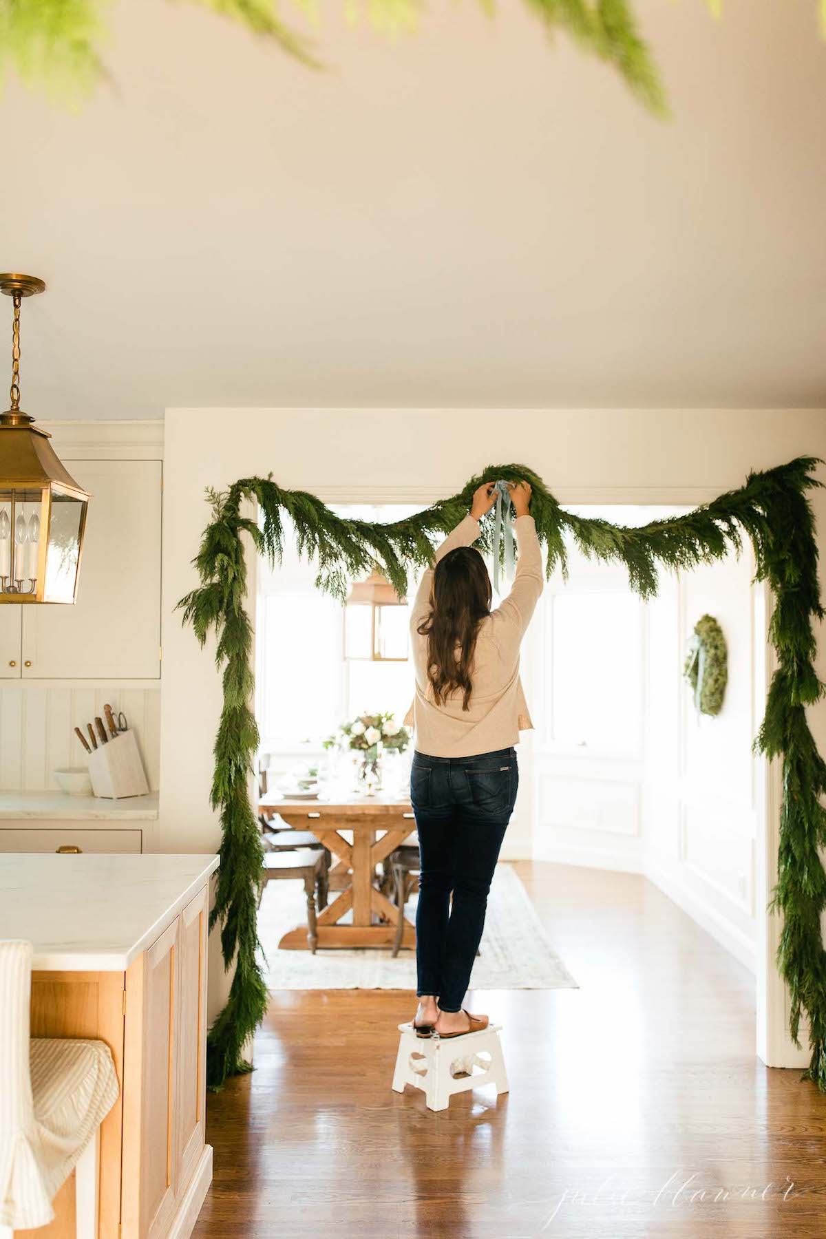 A woman is carefully hanging a blue Christmas garland in her kitchen, adorning her holiday home with festive decorations.