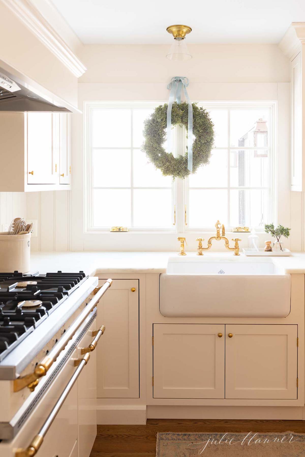 A white kitchen with gold accents and a wreath hanging over the sink, adorned with blue Christmas decorations.
