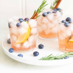 Three glasses of bourbon smash cocktails on a white tray, garnished with blueberries, lemon, rosemary and cinnamon