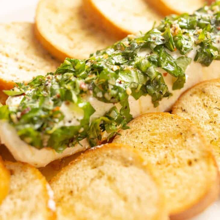 A goat cheese appetizer with a single log of chevre cheese, marinated in herbs and oil, crostini to the side.