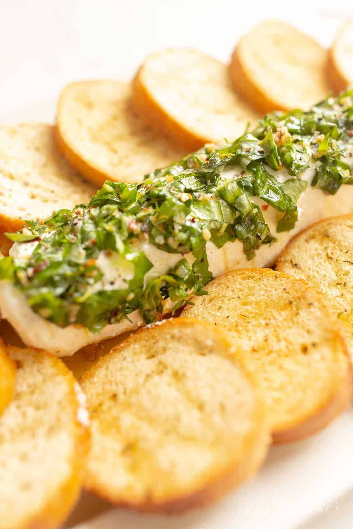 A goat cheese appetizer with a single log of chevre cheese, marinated in herbs and oil, crostini to the side.