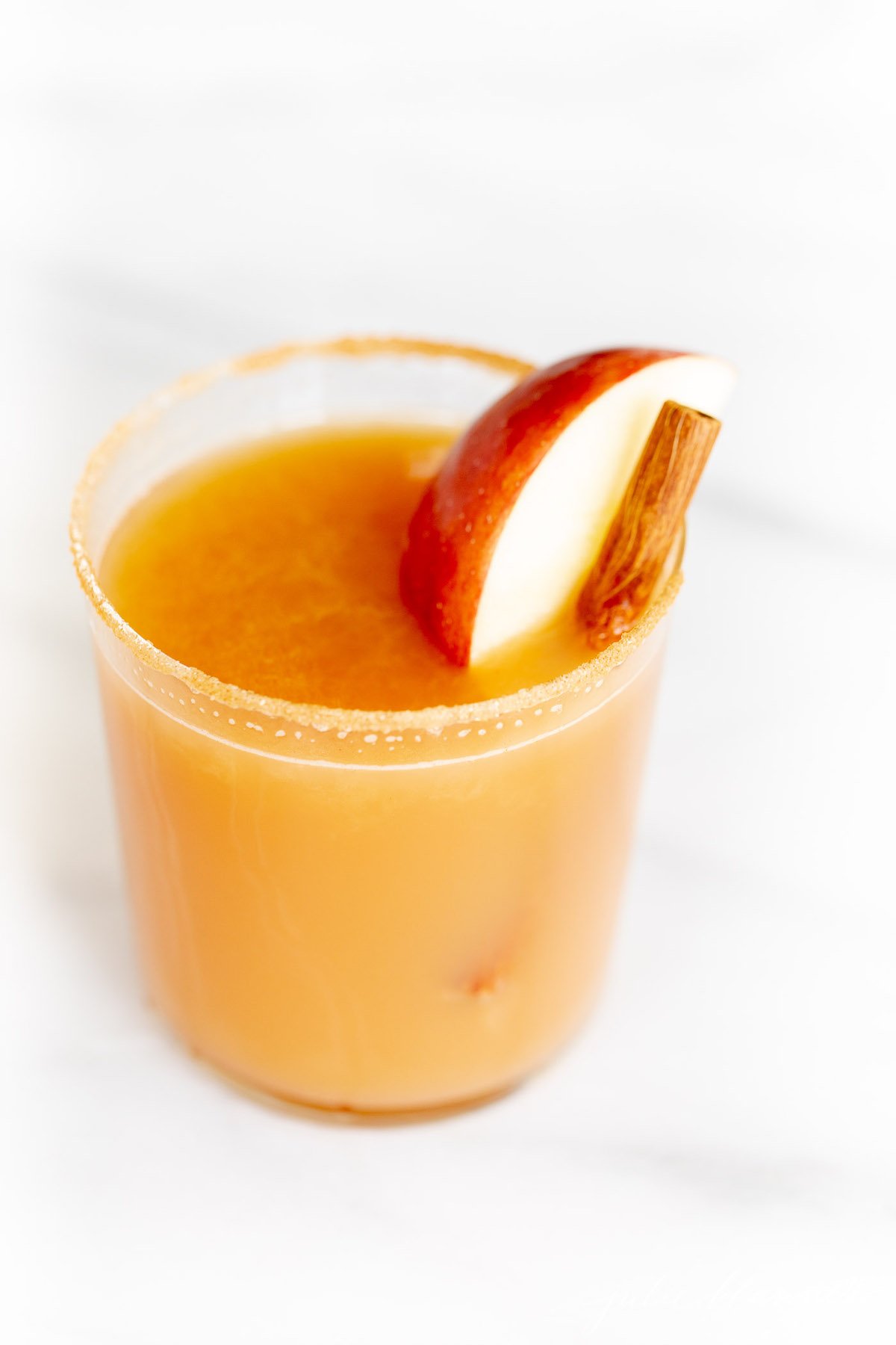 Cinnamon toast cocktail in a glass on marble surface, garnished with apple slice and cinnamon sticks.