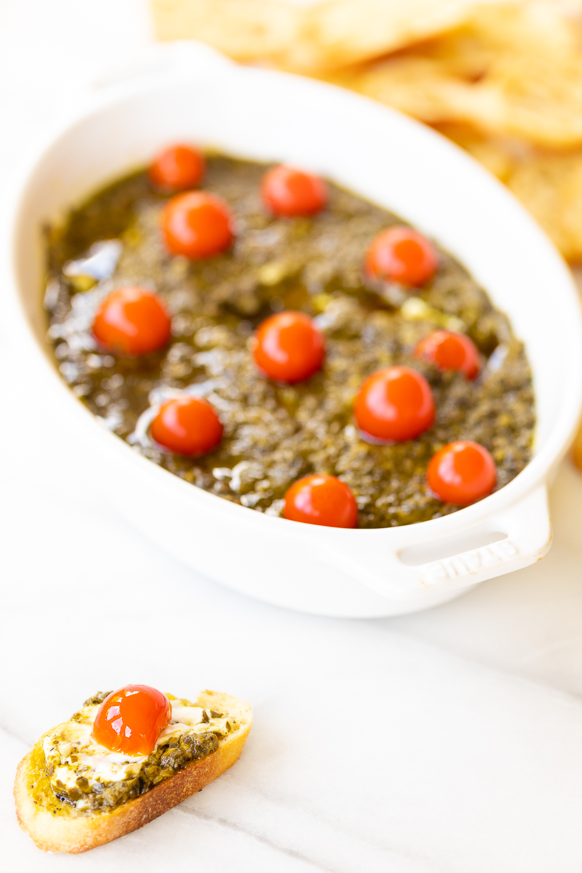 A white bowl of pesto dip with tomatoes and crackers.