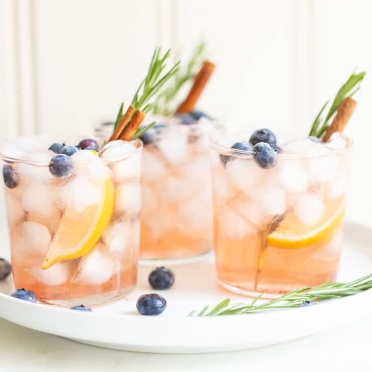 Three Smash cocktails garnished with rosemary, blueberries, oranges, and cinnamon sticks all on a white plate.