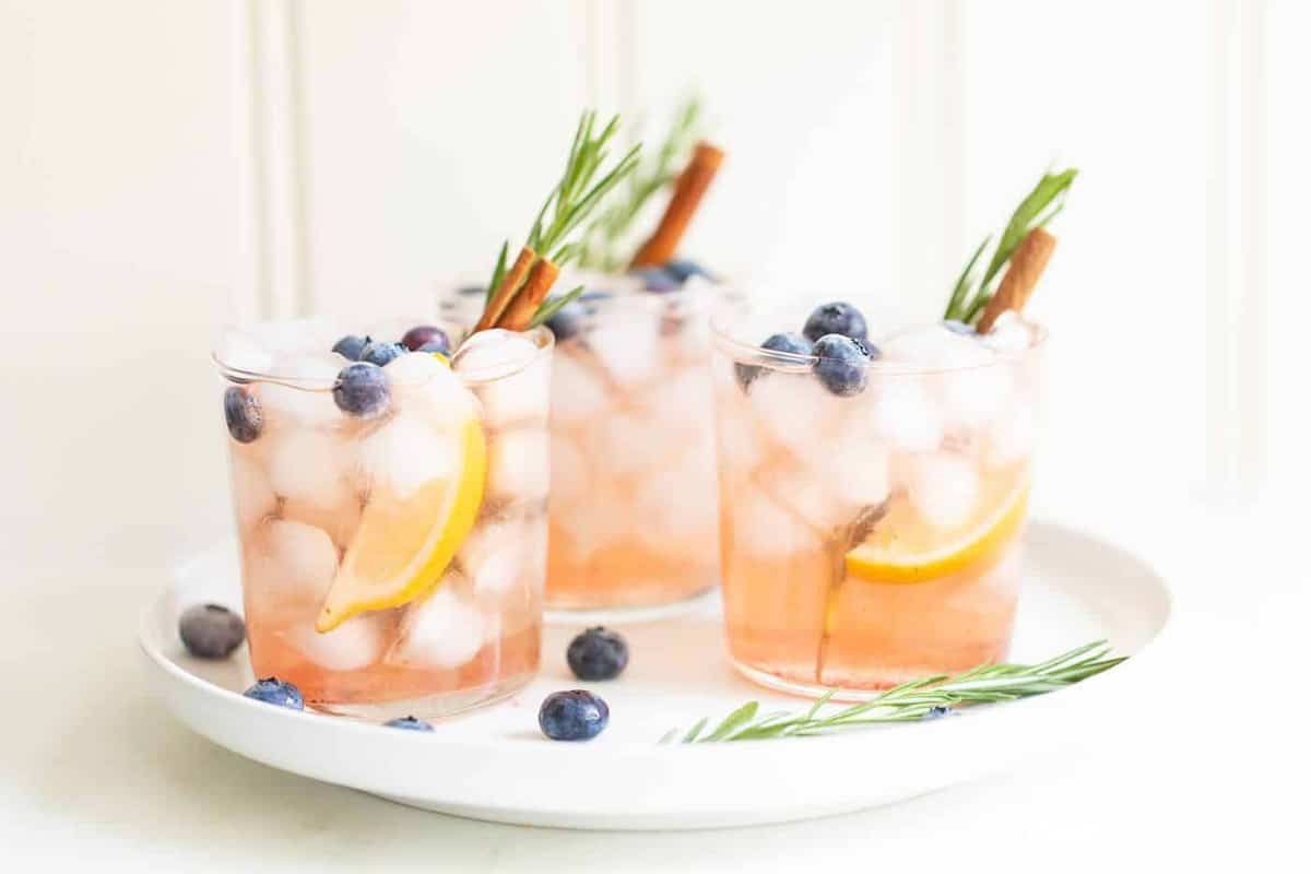 Three Smash cocktails garnished with rosemary, blueberries, oranges, and cinnamon sticks all on a white plate. 