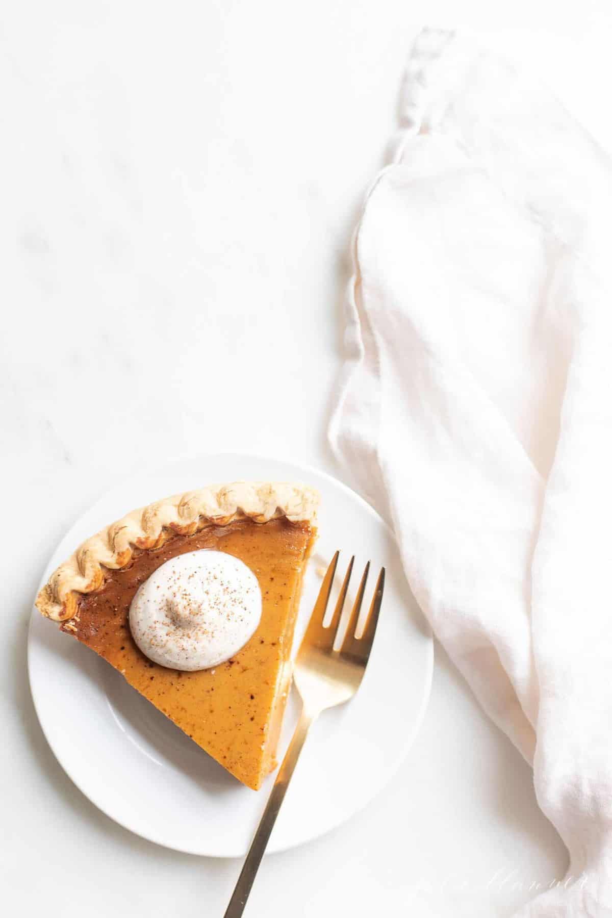 Pumpkin pie topped with egg nog whipped cream on a white plate. A gold fork and napkin are to the side.
