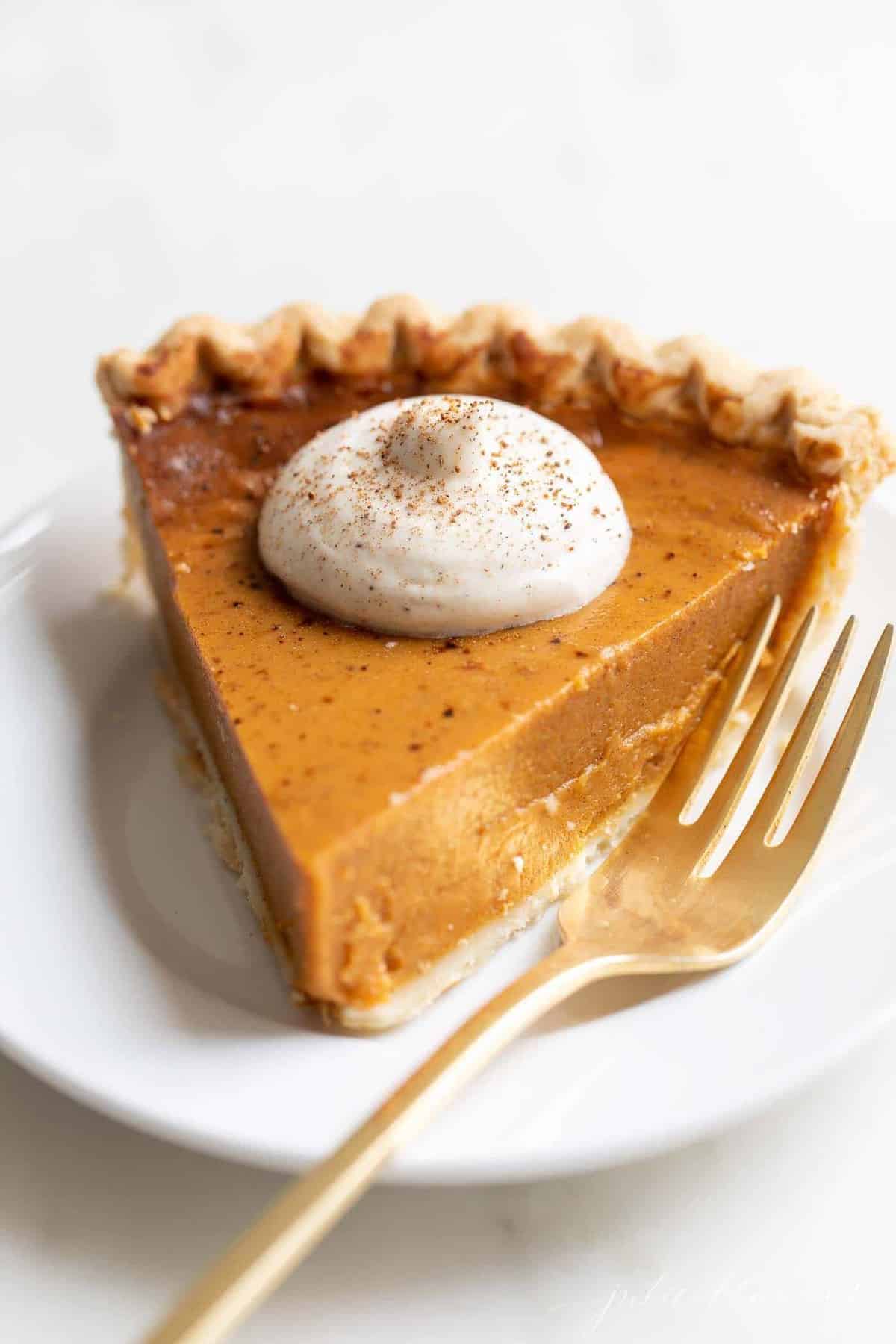 Pumpkin pie topped with egg nog whipped cream on a white plate. A gold fork is to the side.