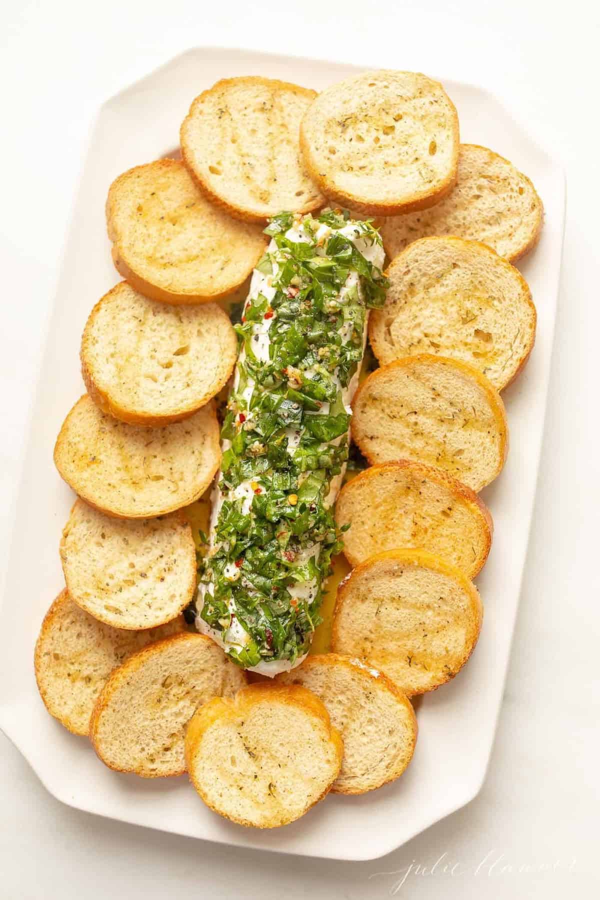 A goat cheese appetizer with a single log of chevre cheese, marinated in herbs and oil, crostini to the side.