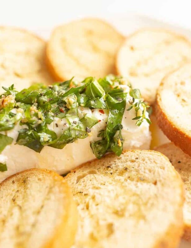 A goat cheese appetizer with a single log of chevre cheese, marinated in herbs and oil, crostini to the side.