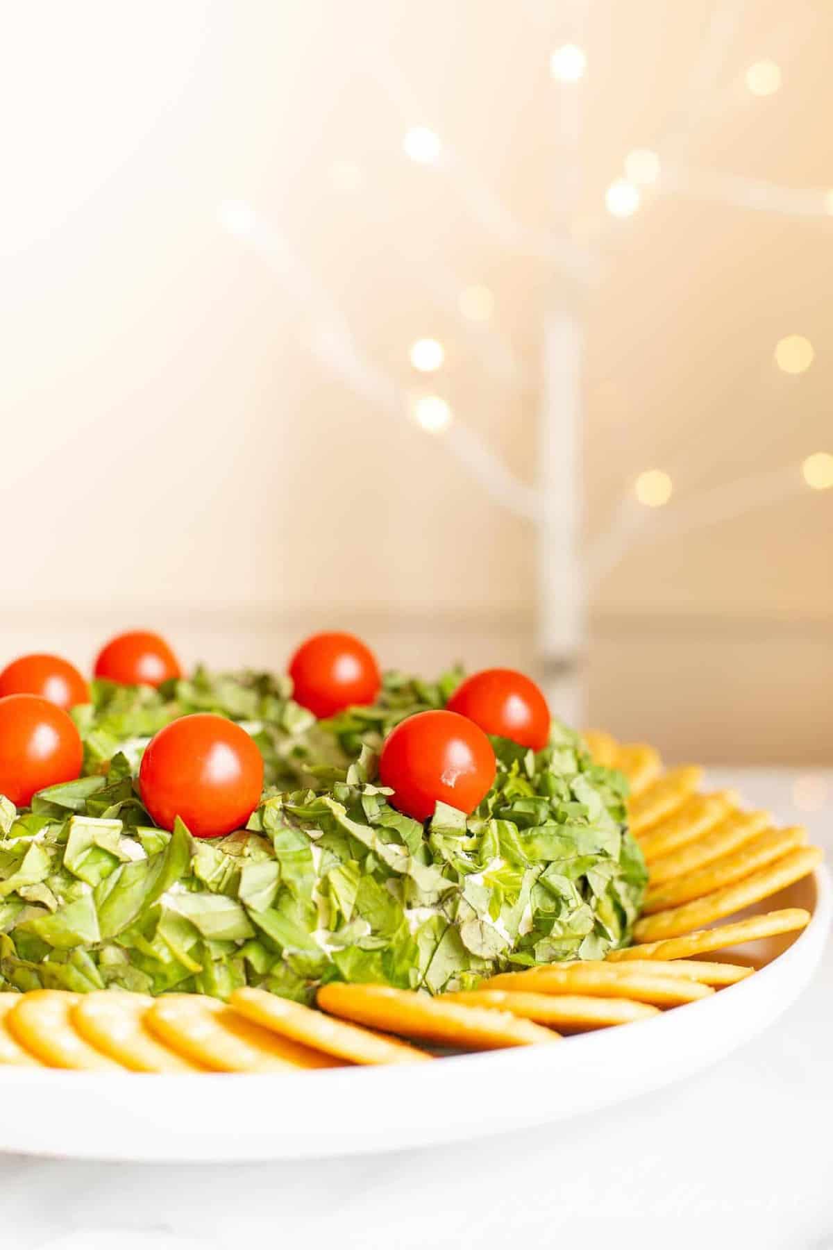A pesto cheeseball Christmas wreath appetizer surrounded by crackers on a platter.