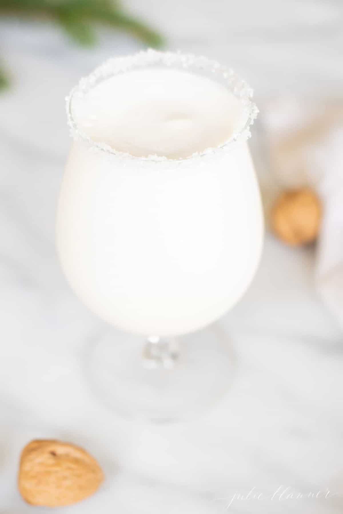 Marble surface with a clear glass filled with a snowball cocktail, rimmed in sugar, with chestnuts tossed around.