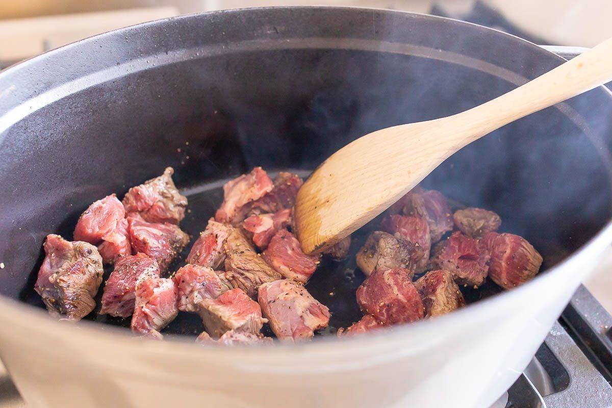 A white cast iron pot on a stove top, wooden spoon stirring chunks of beef as they brown.