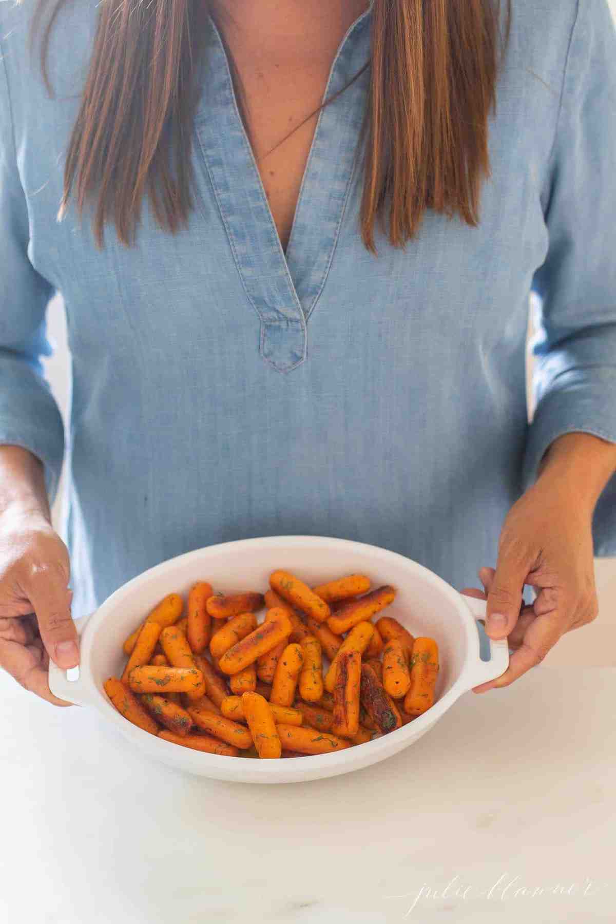 Woman in blue dress holding an oval baking dish filled with braised carrots. 